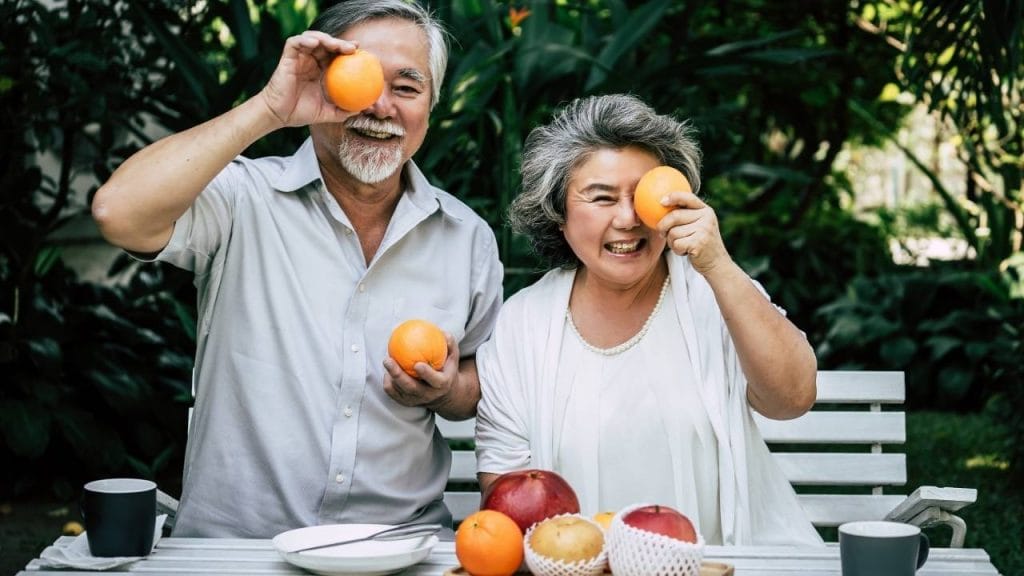 Homem e e mulher de meia idade, segurando frutas em frente aos seus rostos. Sentados atrás de uma mesa com frutas, pratos e canecas, demonstrando uma alimentação balanceada para prevenção dos problemas articulares.