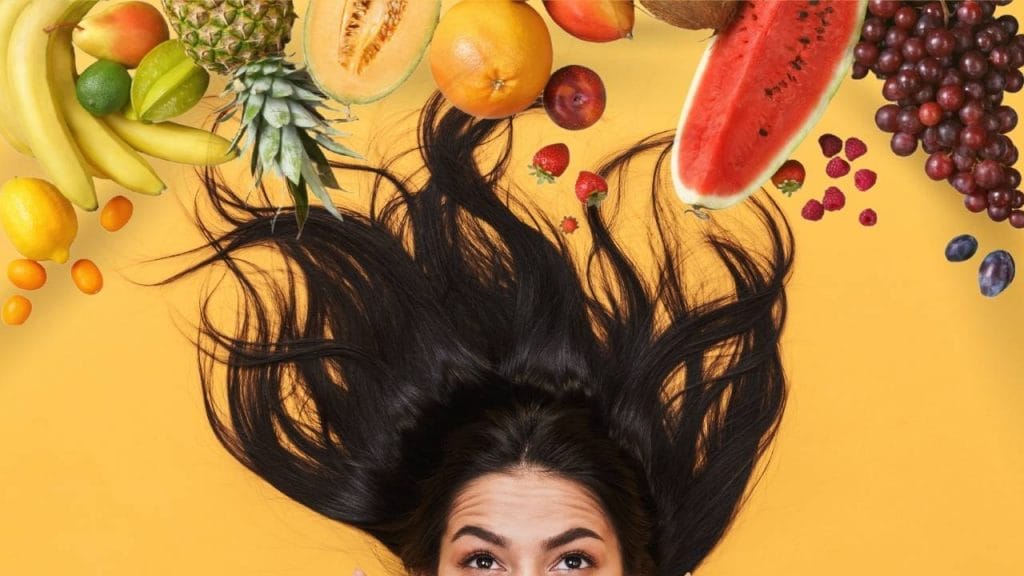 Mulher deitada com os cabelos espalhados no chão e acima dela, diversas frutas.