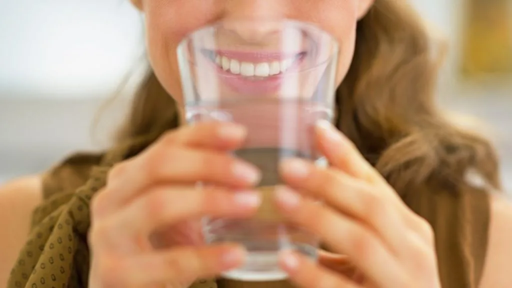 Na imagem, uma mulher leva em direção a sua boca, um copo de água com as duas mãos.