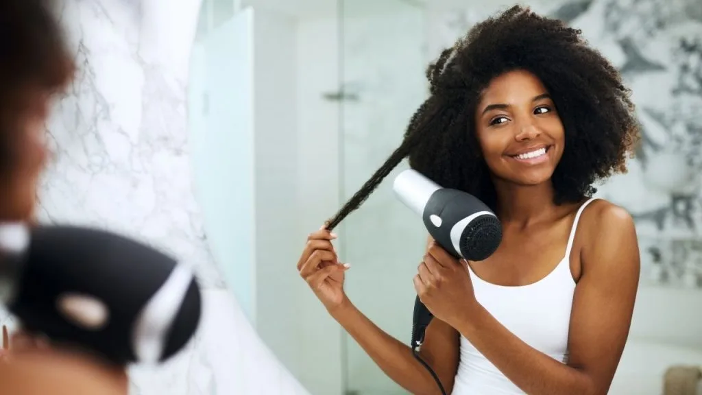 Uma mulher em frente ao espelho, segurando uma mecha com a uma das mãos e com a outra o secador de cabelos.