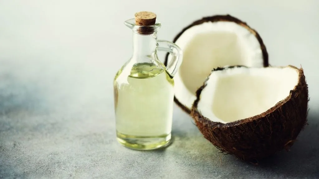 Na imagem, um coco partido ao meio, com seu conteúdo a mostra e uma garrafa de vidro contendo o óleo da fruta.