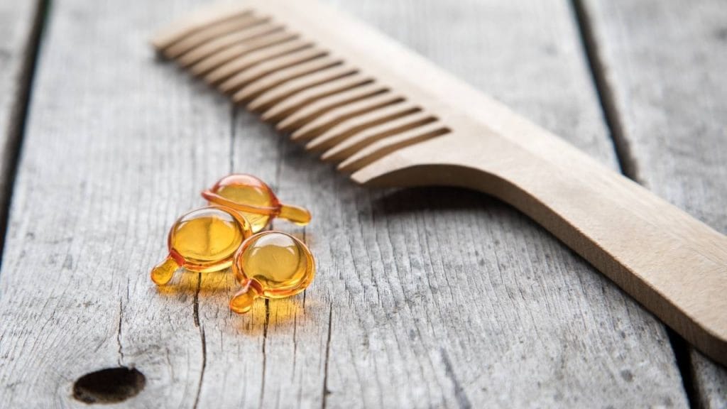 Imagem com um pente de madeira e três capsulas de suplemento de Omega sob uma bancada de madeira.