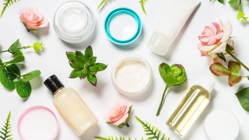 Em uma mesa, está disposta algumas folhas e flores e frascos de cosméticos como cremes, hidratantes, água de limpeza e loções.