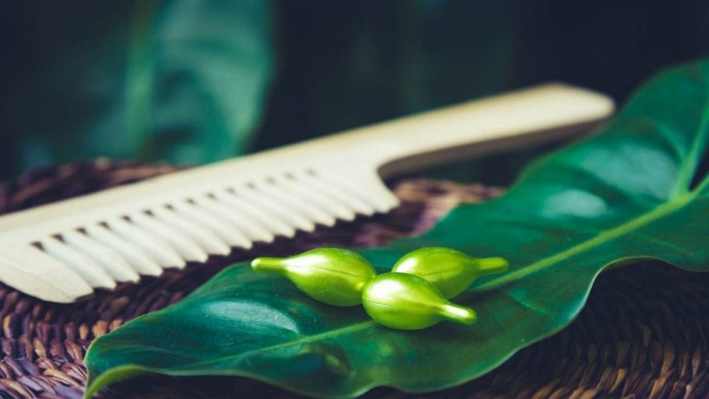 Na imagem, um pente de cabelo, junto a três capsulas de Vitamina E sob uma folha.