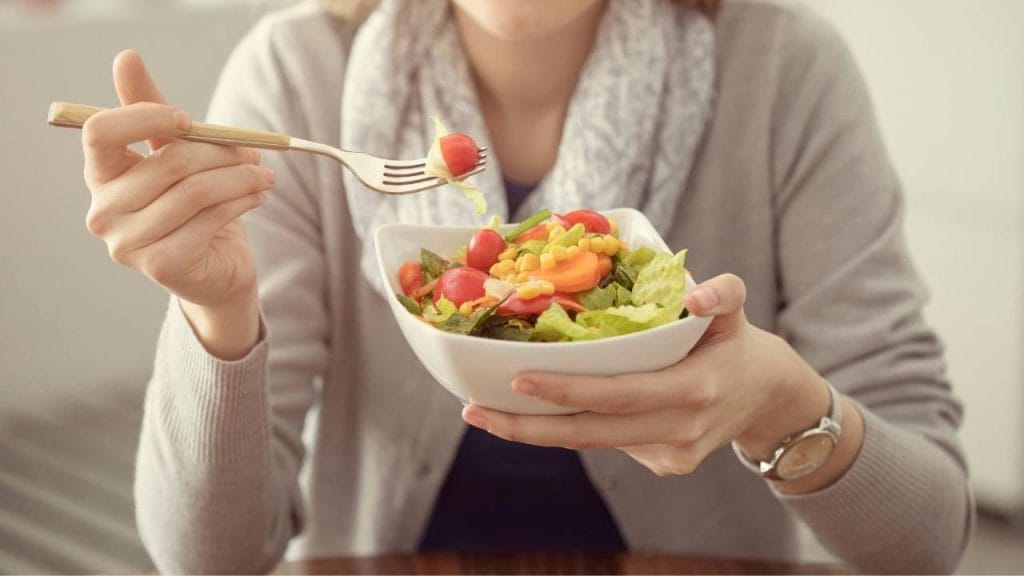Mulher se alimentando adequadamente, com uma pequena travessa contendo verduras e legumes.